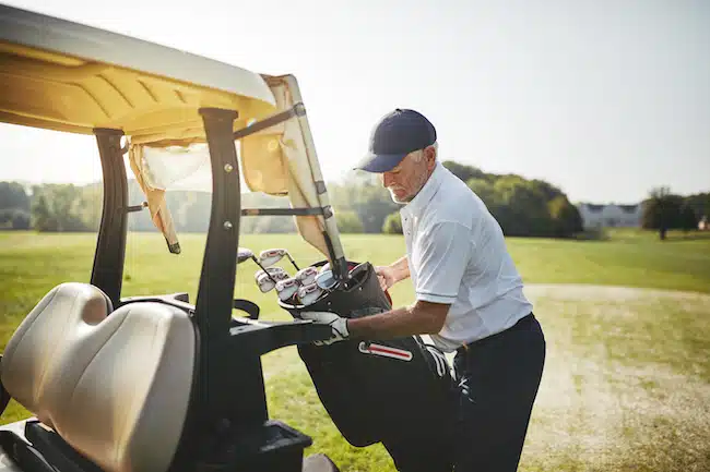 Senior man placing his bag full of clubs on a cart while playing a round of golf on a sunny day