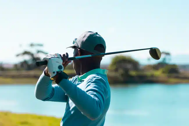 Side view of african american young man taking golf shot with club against lake and clear sky. summer, copy space, golf, cap, unaltered, nature, sport and weekend activities concept.