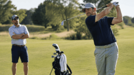A weekend amateur golfer mid-swing on a sunlit fairway, playing partner watching in background with arms crossed