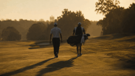 A lone professional golfer photographed from behind in late afternoon golden-hour light, walking down a fairway with his caddie