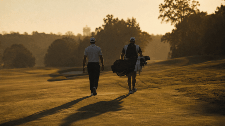 A lone professional golfer photographed from behind in late afternoon golden-hour light, walking down a fairway with his caddie
